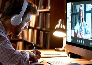 Estudiante varón con auriculares videoconferencia, viendo webinar, clase de formación en línea, reunión de chat virtual con profesor o entrenador a distancia usando el ordenador, tomando notas.