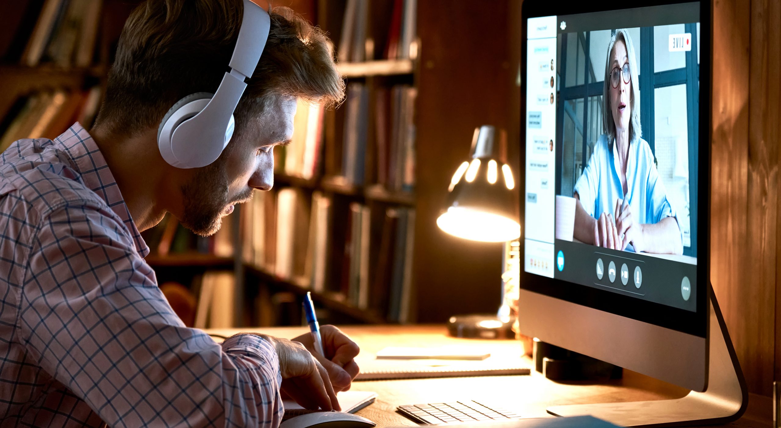 Estudiante varón con auriculares videoconferencia, viendo webinar, clase de formación en línea, reunión de chat virtual con profesor o entrenador a distancia usando el ordenador, tomando notas.